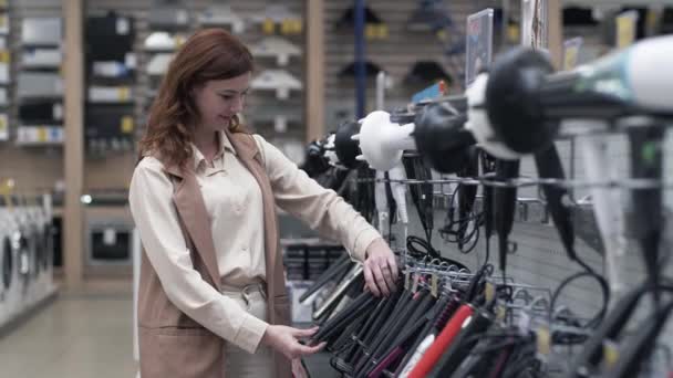 Chica comprador elige estiladores de pelo en los estantes de una tienda de artículos eléctricos — Vídeo de stock