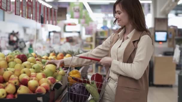 Alimentación saludable, ama de casa bonita con cesta de comestibles selecciona en los estantes del supermercado verduras y frutas respetuosas del medio ambiente — Vídeo de stock