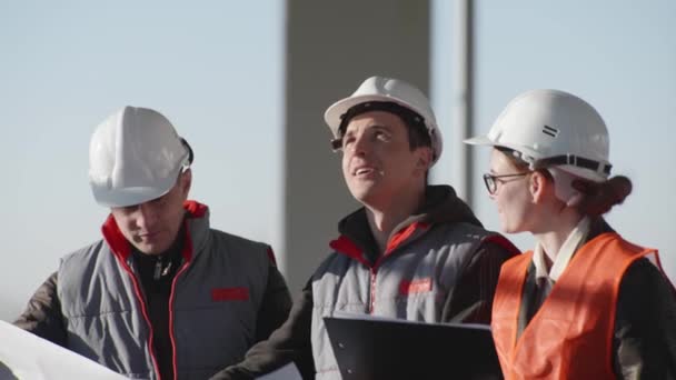 Розробка, дружня команда інженерів та архітекторів у захисних касках обговорюють креслення на будівельному майданчику — стокове відео