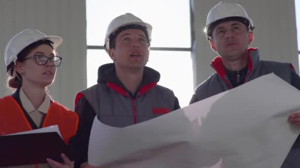 Team von männlichen und weiblichen Baufachleuten in Schutzhelmen mit Zeichnungen in den Händen inspiziert den Bau der überdachten Halle — Stockvideo
