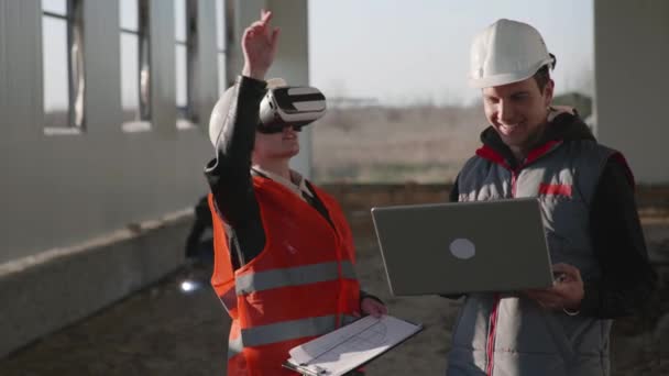 Modern teknik, kvinnlig och manlig ingenjör i viar glasögon och laptop i hand i skyddshjälmar diskutera ny byggnad — Stockvideo