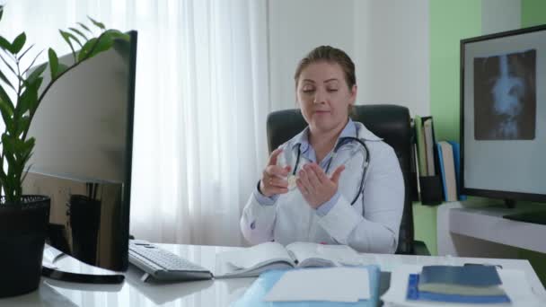 A fonendoszkópos női orvos beszámol a betegnek a higiéniai és biztonsági előírásokról, bemutatja, hogyan kell használni a fertőtlenítőt a kamerán, miközben az orvosi irodában ül, — Stock videók