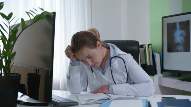 Втомлена жінка лікар тримає голову в руці, відчуває себе погано через хворобу або втому, працює за комп'ютером і дивиться на монітор, сидячи в медичній кімнаті — стокове відео