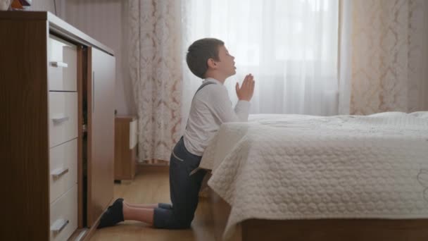 Religie, kleine christelijke jongen met geloof en hoop in het hart met gesloten ogen gebalde handen bidt tot God op zijn knieën in de kamer in de buurt van bed — Stockvideo