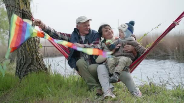 Niñez, niño feliz sentado en brazos y abuelos amorosos en hamaca y jugando con una cometa — Vídeos de Stock