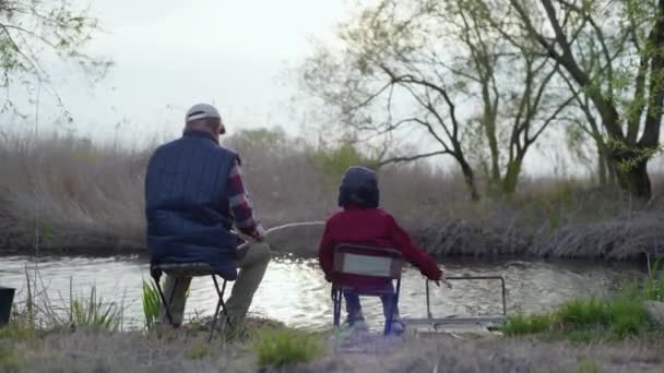 Старый рыбак вместе со своим счастливым внуком ловит рыбу на удочке у реки — стоковое видео