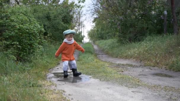 Divertida infancia, niño varón feliz en sombrero y botas de goma saltando alegremente en charco — Vídeos de Stock