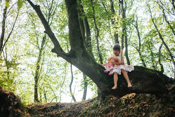 Niña en un vestido con muñeca se sienta en un árbol en el bosque — Foto de Stock