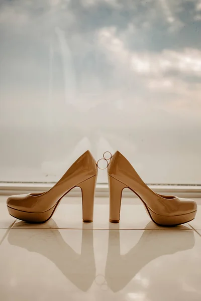 Anillos de boda de oro entre un par de zapatos blancos de tacón alto. Detalles de la boda en el piso del espejo cerca de ventana panorámica — Foto de Stock