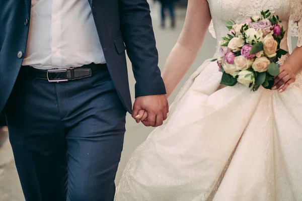 Bouquet de mariage entre les mains des mariés. Couple de mariage marche se tenant la main. En robe blanche et veste bleue — Photo