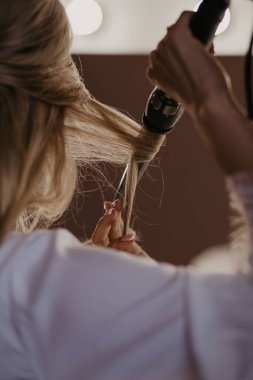 Gelin evde saç maşasıyla saçını kıvırıyor. Gelinin hazırlıkları. Kapatın. Kadınlar saç stili yapıyor ve uzun kıvırcık saçlarını düzleştiriyor. Güzellik ve saç bakım konsepti