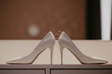 Bir çift yüksek topuklu ayakkabının arasında 2 altın alyans. Düğün detayları aynanın yanındaki masada.