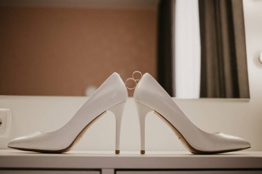 Bir çift yüksek topuklu ayakkabının arasında 2 altın alyans. Düğün detayları aynanın yanındaki masada.