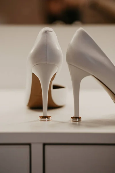 2 Two Gold wedding rings under pair of white high heel shoes. Wedding details on white table near mirror — Stock Photo, Image