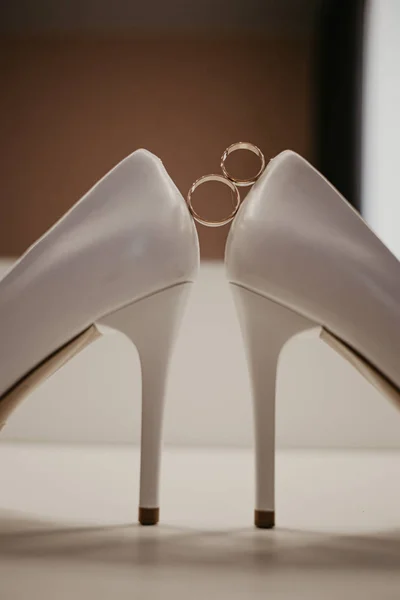 Anillos de boda de oro entre un par de zapatos blancos de tacón alto. Detalles de la boda en la mesa cerca del espejo — Foto de Stock