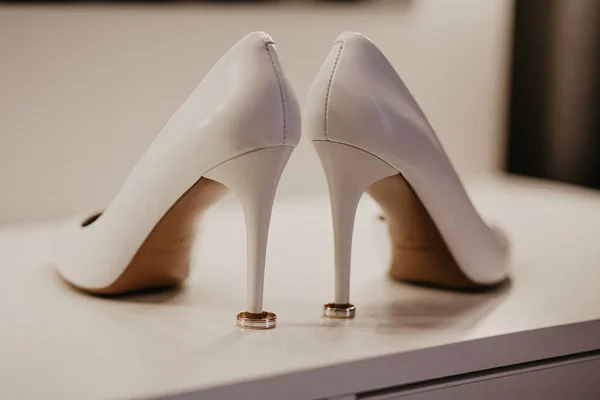 2 Dos anillos de boda de oro bajo un par de zapatos blancos de tacón alto. Detalles de la boda en mesa blanca cerca del espejo — Foto de Stock