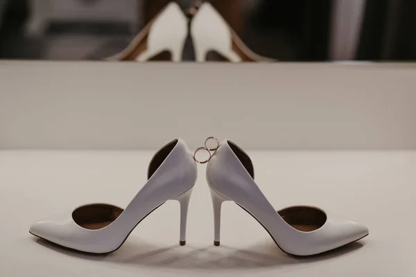 Anillos de boda de oro entre un par de zapatos blancos de tacón alto. Detalles de la boda en la mesa cerca del espejo — Foto de Stock