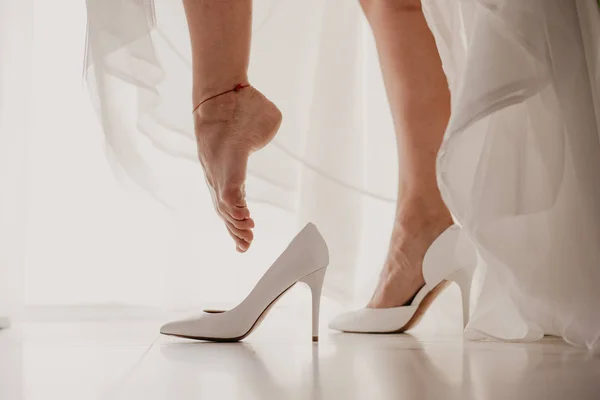 Pengantin wanita mengenakan sepatu hak tinggi di lantai cermin dekat jendela panorama pada hari pernikahan. Mempelai meletakkan kaki elegan nya ke sepatu putih — Foto Stok Gratis