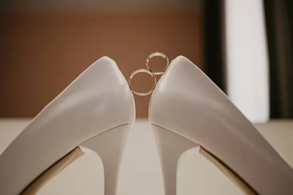 2 Dua cincin kawin emas antara sepasang sepatu hak tinggi putih. Detail pernikahan di meja dekat cermin — Stok Foto