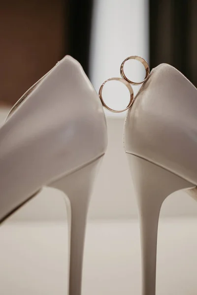 2 Dos anillos de boda de oro entre un par de zapatos blancos de tacón alto. Detalles de la boda en la mesa cerca del espejo — Foto de Stock