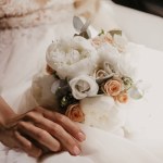 Bröllopsbukett i händerna på bruden. Vacker brud sitter och håller bröllop bukett med vackra rosor och pioner. I vit klänning och fin monicure. Närbild