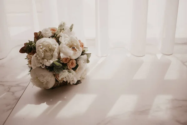 Wedding bouquet with beautiful roses and pions with 2 wedding rings lying on the floor in front of window with curtains. Long shadow of from window — Stock Photo, Image