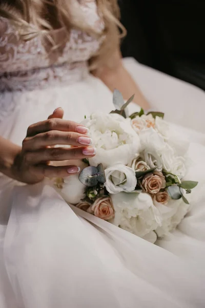Wedding bouquet in hands of bride. Beautiful bride sitting and touching wedding bouquet with beautiful roses and pions. In white dress and nice monicure. Close up — Stock Photo, Image