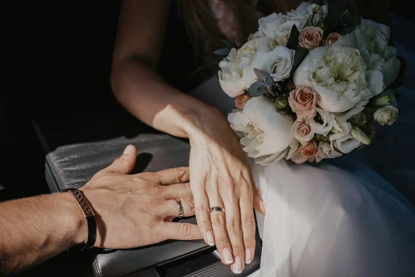 Uma mão de casal de casamentos tocam-se. Mãos recém-casados com anéis de casamento de perto. Mãos nupciais com anéis no braço de couro preto em assentos de carro. Sombra por causa do sol — Fotografia de Stock