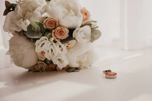 Buquê de casamento com belas rosas e piões com 2 alianças de casamento deitado no chão perto da janela com cortinas. Longa sombra da janela — Fotografia de Stock Grátis