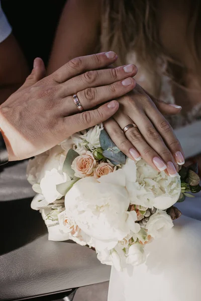 A hand of wedding couple touch each other over wedding bouquet. Hands newlyweds with wedding rings close up. Bridal hands with rings on black leather armrest on car seats. Shadow because of the sun — Stock Photo, Image