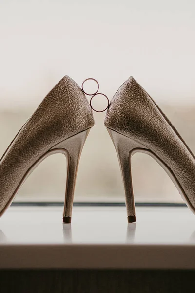 Золотые обручальные кольца между парой туфель на высоком каблуке. Свадебные детали на зеркальном полу возле панорамного окна. Закрыть — стоковое фото
