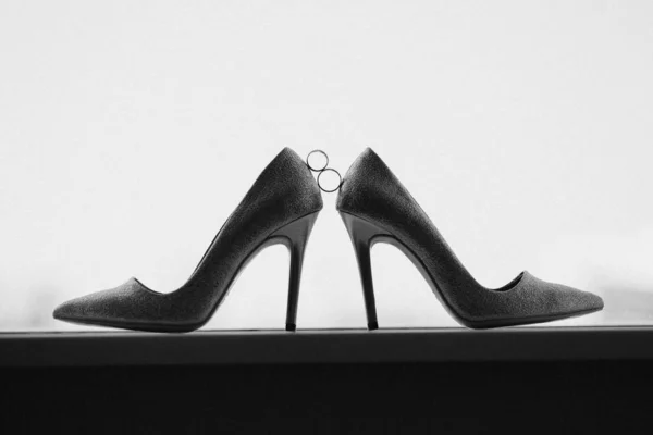 Золотые обручальные кольца между парой туфель на высоком каблуке. Свадебные детали на зеркальном полу возле панорамного окна. Монохромный. Черное и белое — стоковое фото