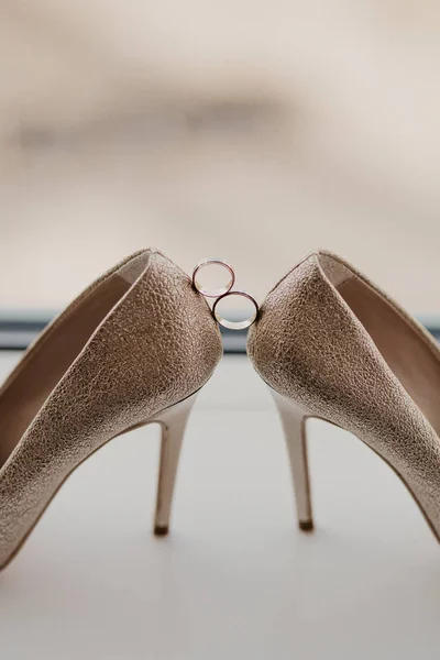 Anillos de boda de oro entre un par de zapatos de tacón alto de oro. Detalles de la boda en el piso del espejo cerca de ventana panorámica. De cerca. — Foto de Stock