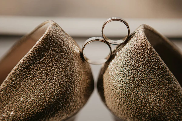 Anéis de casamento de ouro entre o par de sapatos de salto alto dourado. Detalhes do casamento no chão espelho perto da janela panorâmica. Fecha. Macro — Fotografia de Stock
