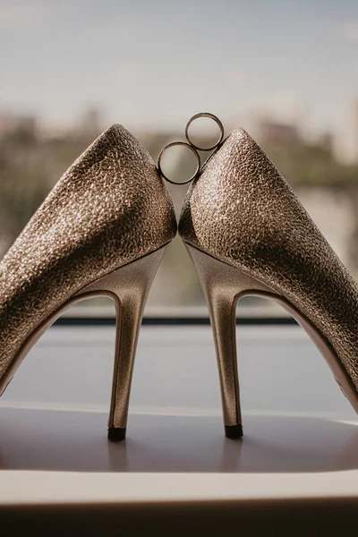 Gold wedding rings between pair of golden high heel shoes. Wedding details on mirror floor near panoramic window. On sunshine — Stock Photo, Image