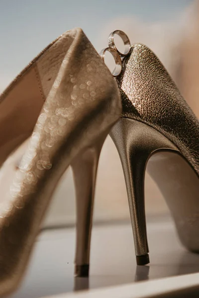 Anillos de boda de oro entre un par de zapatos de tacón alto de oro. Detalles de la boda en el piso del espejo cerca de ventana panorámica. De cerca. Macro. En el sol — Foto de stock gratis