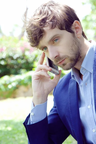 Bonito jovem homem de negócios fazendo uma chamada com seu smartphone fora em um parque verde — Fotografia de Stock