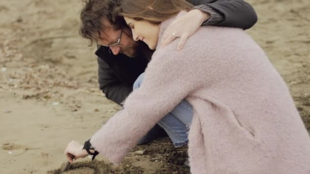 Schöne Frau bemalt Herz auf Sand umarmt von liebenden Freund Nahaufnahme — Stockvideo