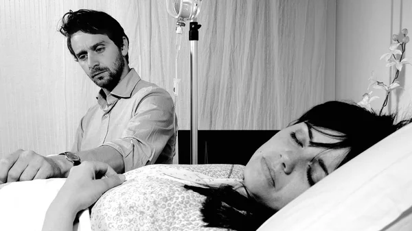 Triste marido preocupado com sua esposa doente que está dormindo em uma cama de hospital preto e branco — Fotografia de Stock