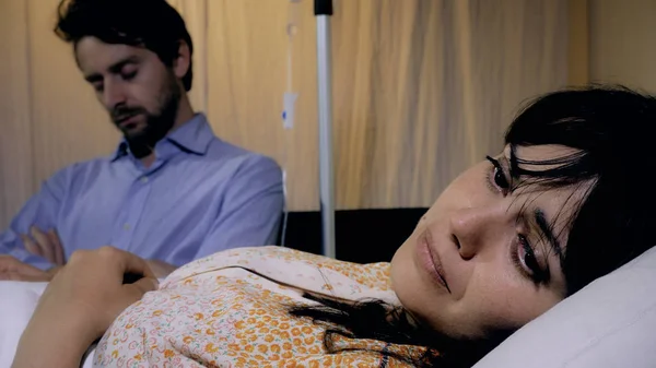 Хвора молода пригнічена жінка в лікарні з чоловіком спить в кріслі поруч з нею — стокове фото