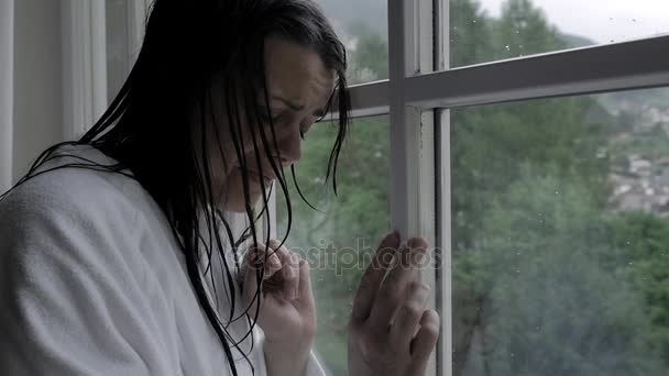 Женщина с мокрыми волосами перед окном с дождём чувствует себя одиноким и подавленным замедленным движением — стоковое видео