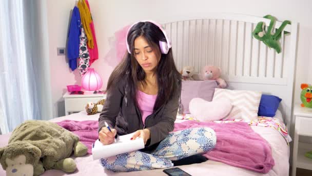 快乐女学生坐在床上听音乐 而做家庭作业跳舞 — 图库视频影像