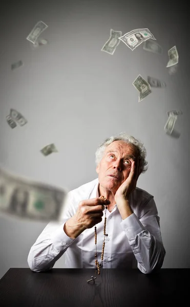 Oude man is bidden met rozenkrans kralen. Dalende dollar biljetten. — Stockfoto