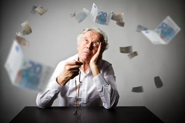 Oude man is bidden met rozenkrans kralen. Dalende waarde van de eurobankbiljetten. — Stockfoto