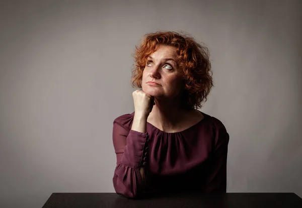 Sad red-haired woman is sitting at the table. Doubts. — Stock Photo, Image