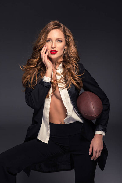 seductive woman touching face with hand and holding american football ball isolated on gray