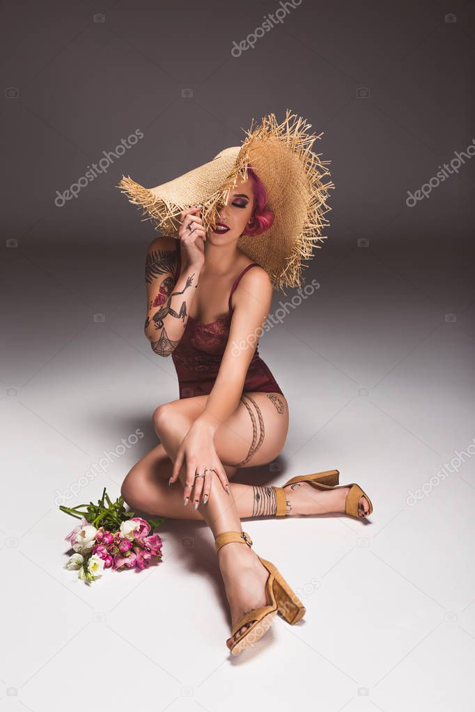 beautiful tattoed pin up girl in lingerie and straw hat with flowers infront of grey background 