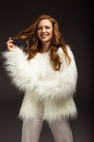 Chica sonriente en chaqueta mullida tocando el pelo aislado en gris - foto de stock