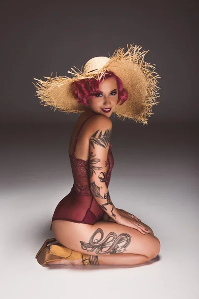 Pretty tattoed pin up girl em lingerie e chapéu de palha sentado no chão na frente de fundo cinza — Fotografia de Stock