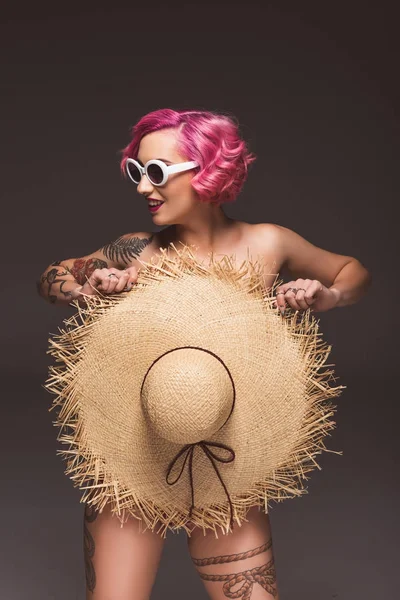 Hermosa mujer de pelo rosa desnuda en gafas de sol que cubren con sombrero de paja delante de fondo gris - foto de stock